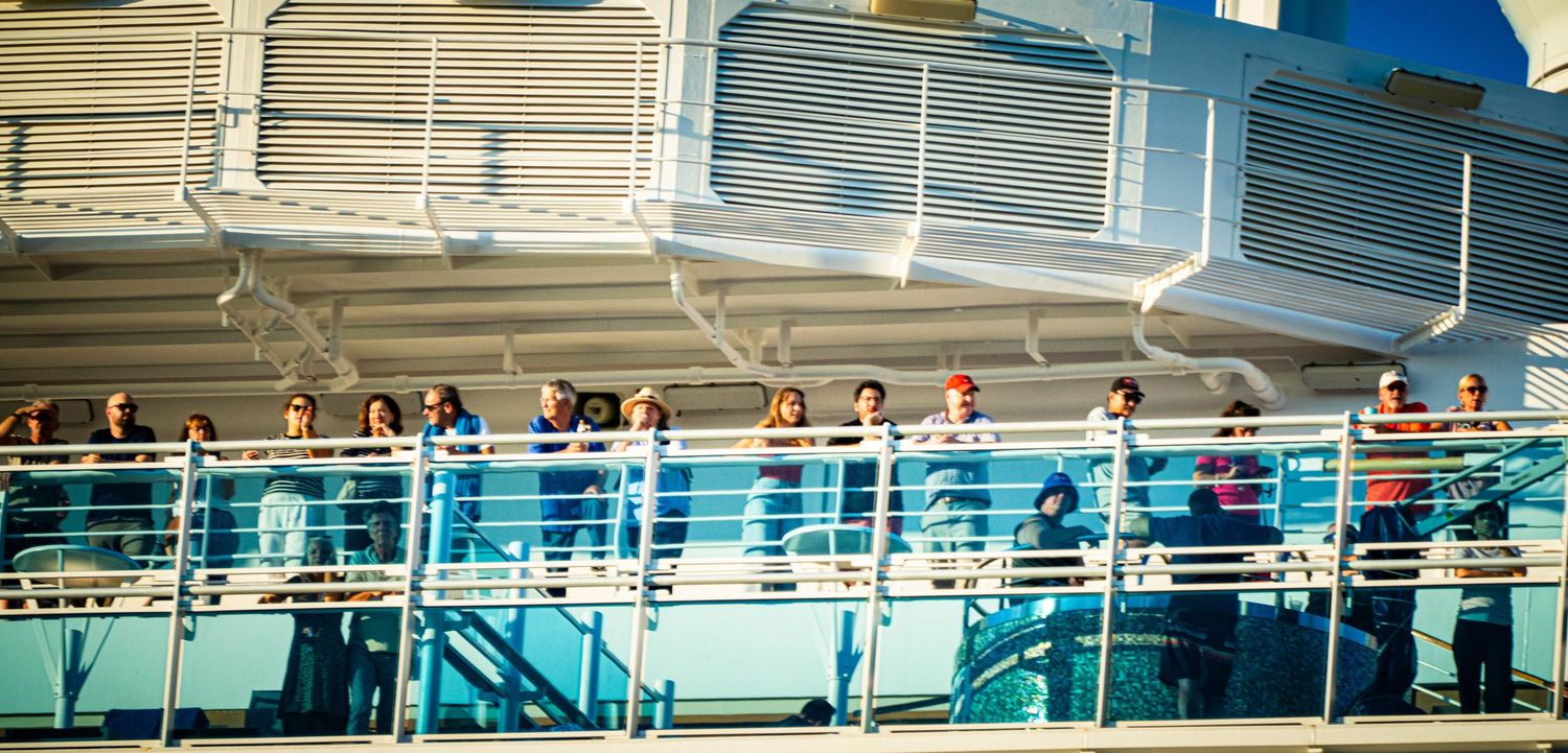 Cruise ship passengers line the outdoor deck railings while watching the sunset over the ocean.