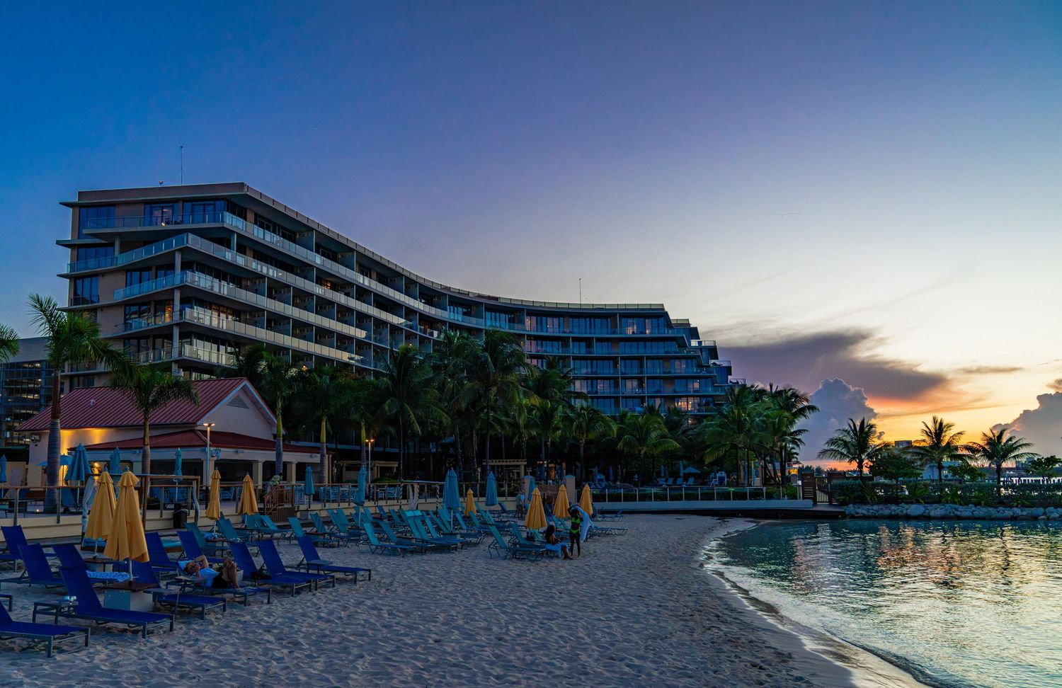 Luxury beachfront resort hotel at sunset with blue lounge chairs, palm trees, and a curved modern facade overlooking calm waters.