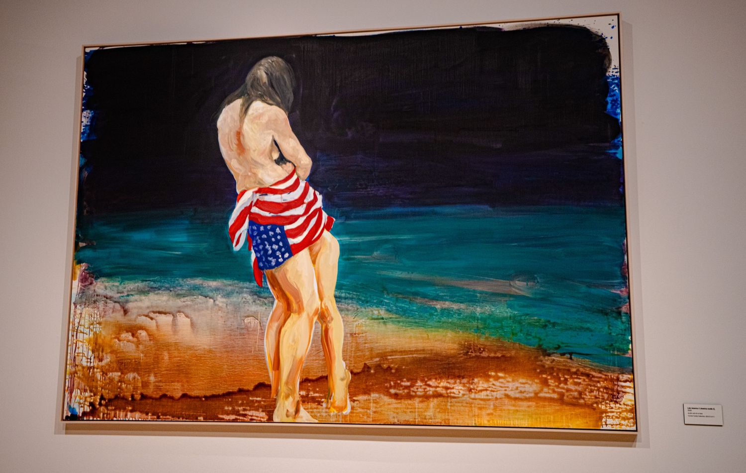 Painting of a woman draped in an American flag standing by a colorful shoreline, displayed in an art gallery.