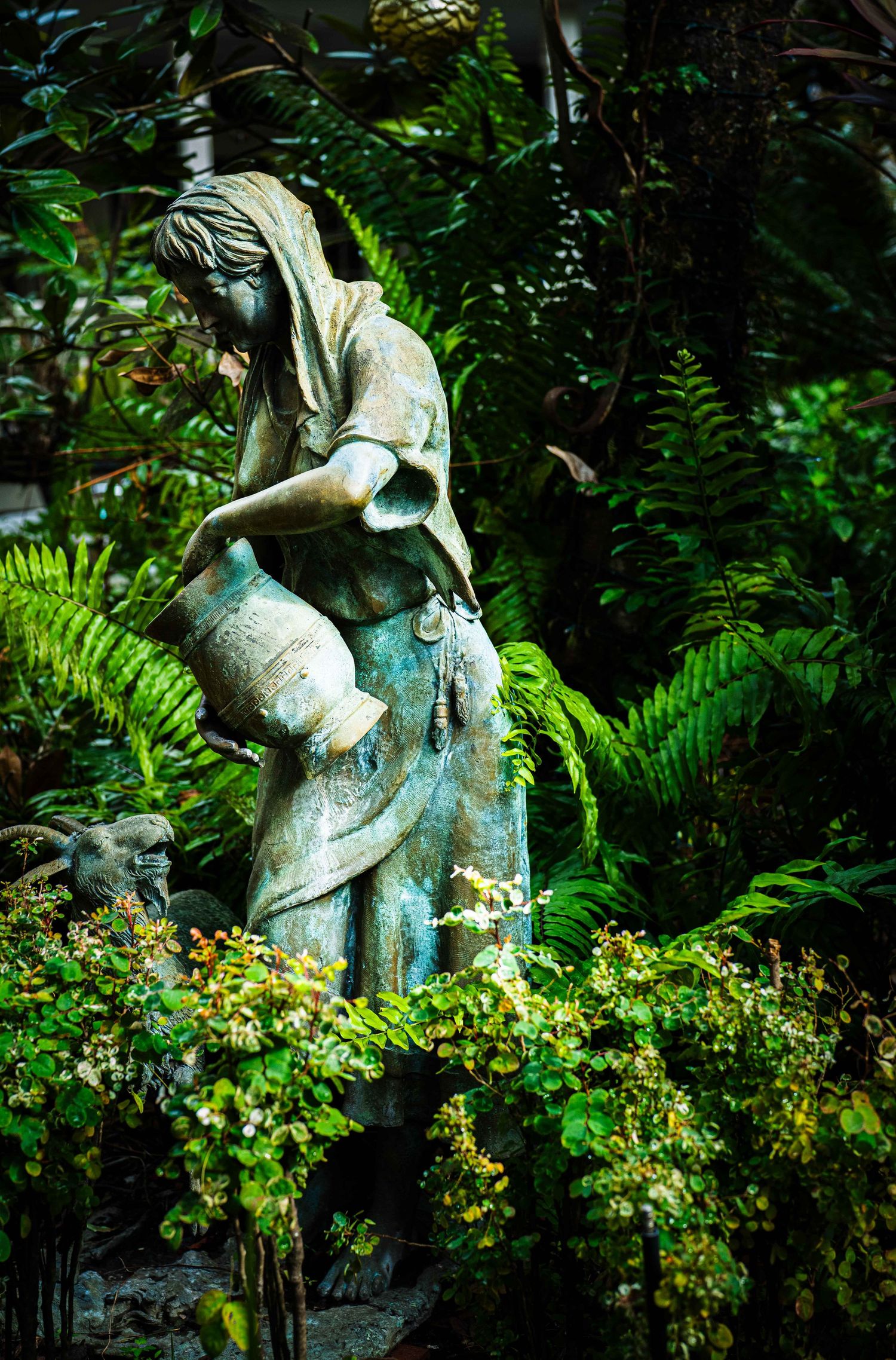 Garden sculpture of children playing on a log surrounded by lush ferns and yellow flowers in a peaceful setting.