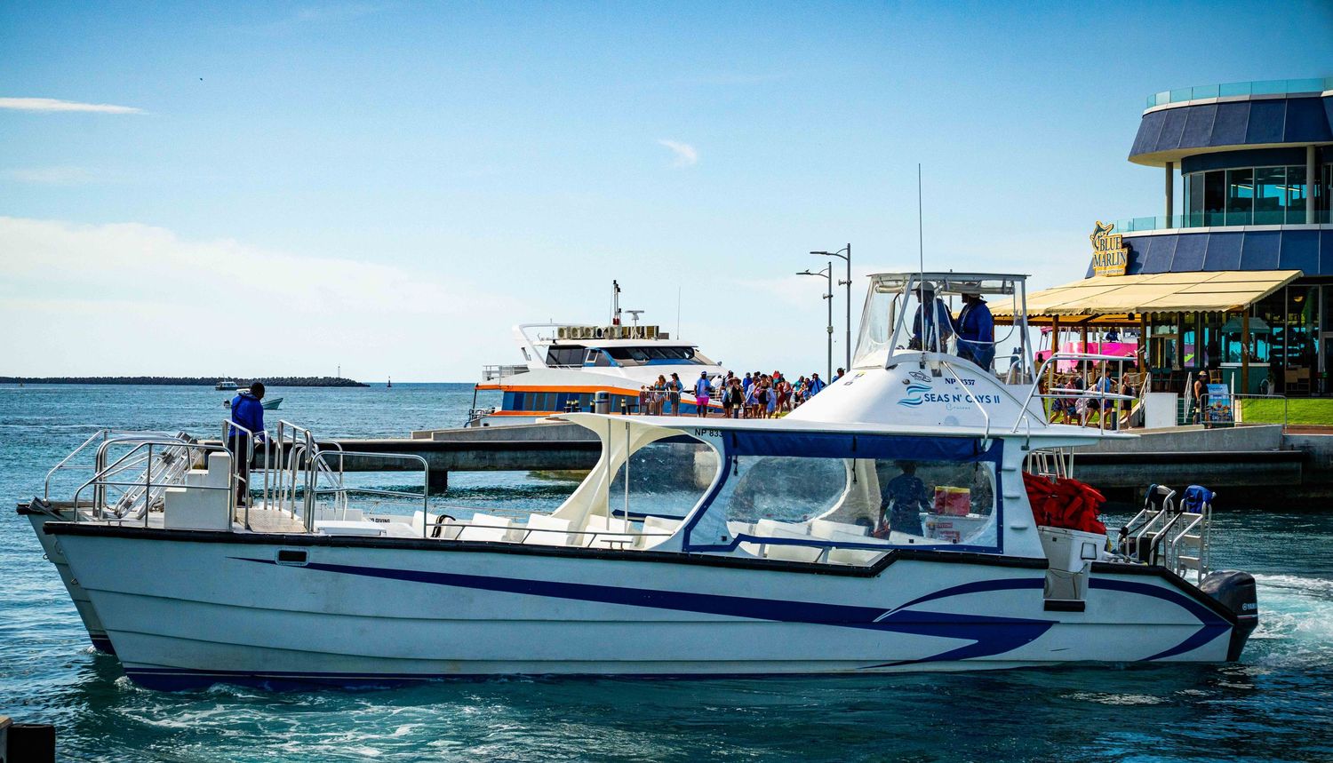 A white passenger ferry boat cruising on blue harbor waters near a busy waterfront dock on a sunny day.