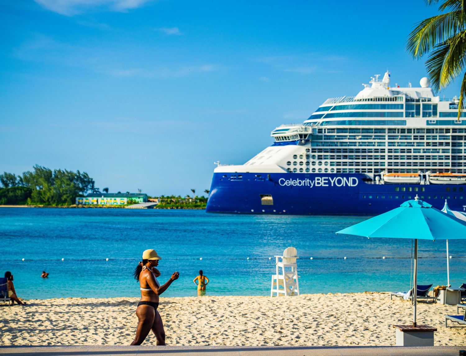 Celebrity Beyond cruise ship docked near a tropical beach with turquoise water, palm trees, and vacationers enjoying the sunny shore.