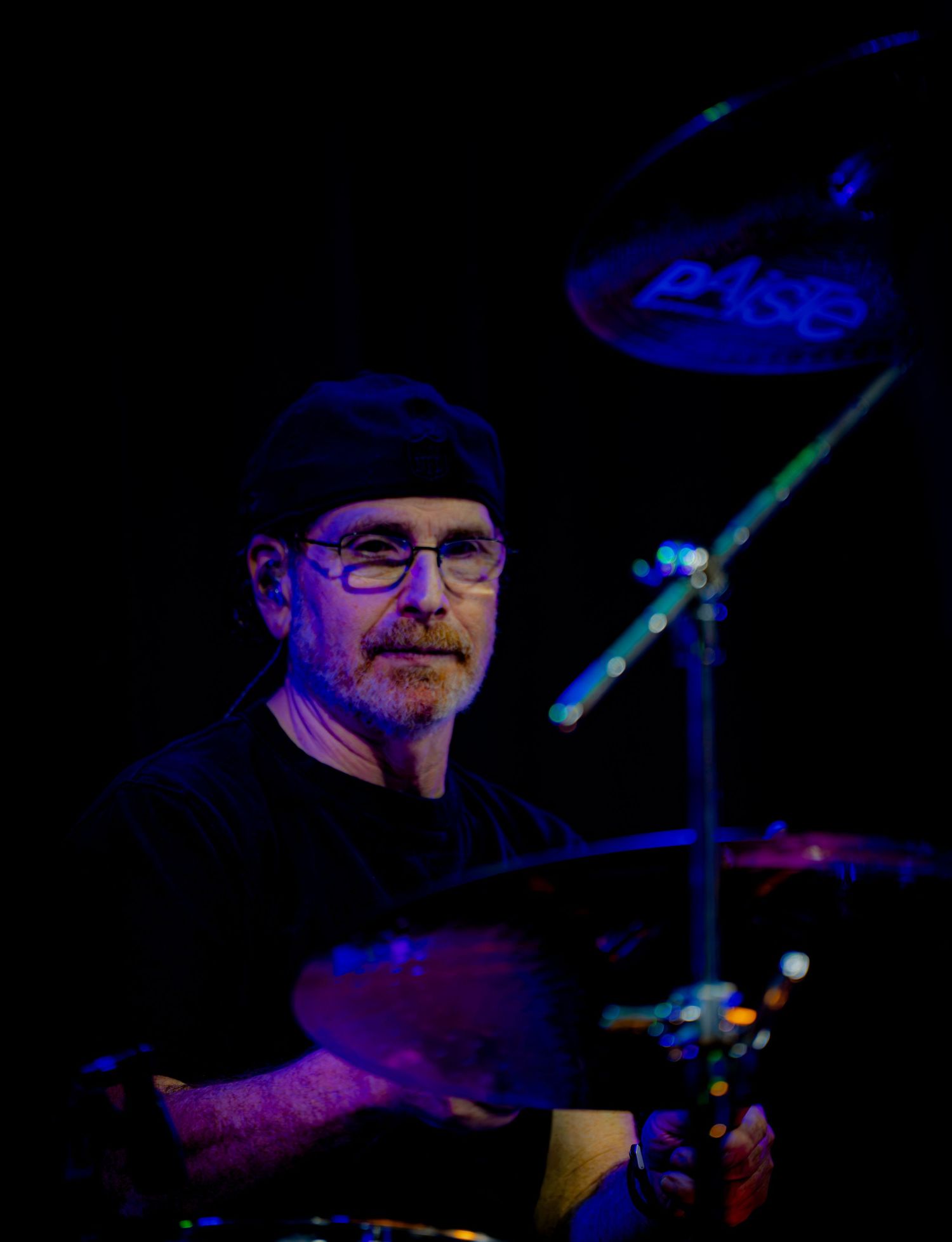 Bearded male drummer wearing glasses and black cap performing on stage with Paiste cymbals in blue stage lighting.