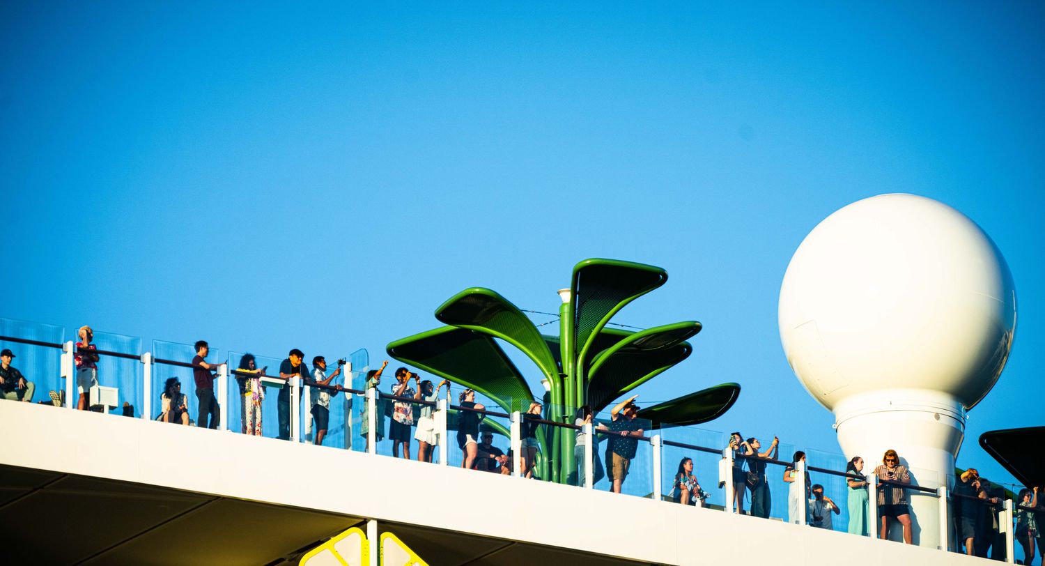 People line up along a railing against bright blue sky with a white sphere and green palm fronds visible.