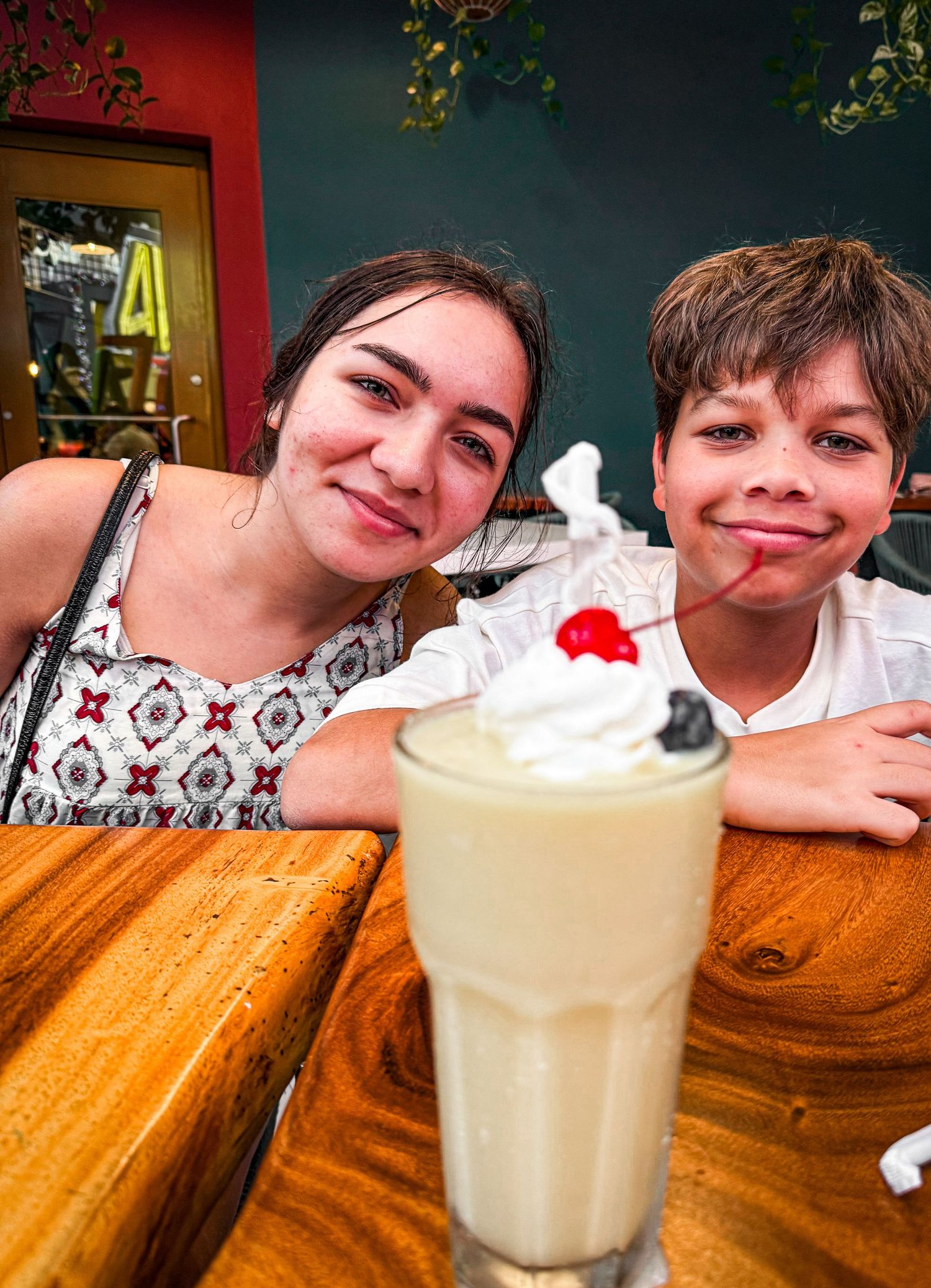 A milkshake topped with whipped cream and a cherry sits on a wooden table with someone resting beside it.