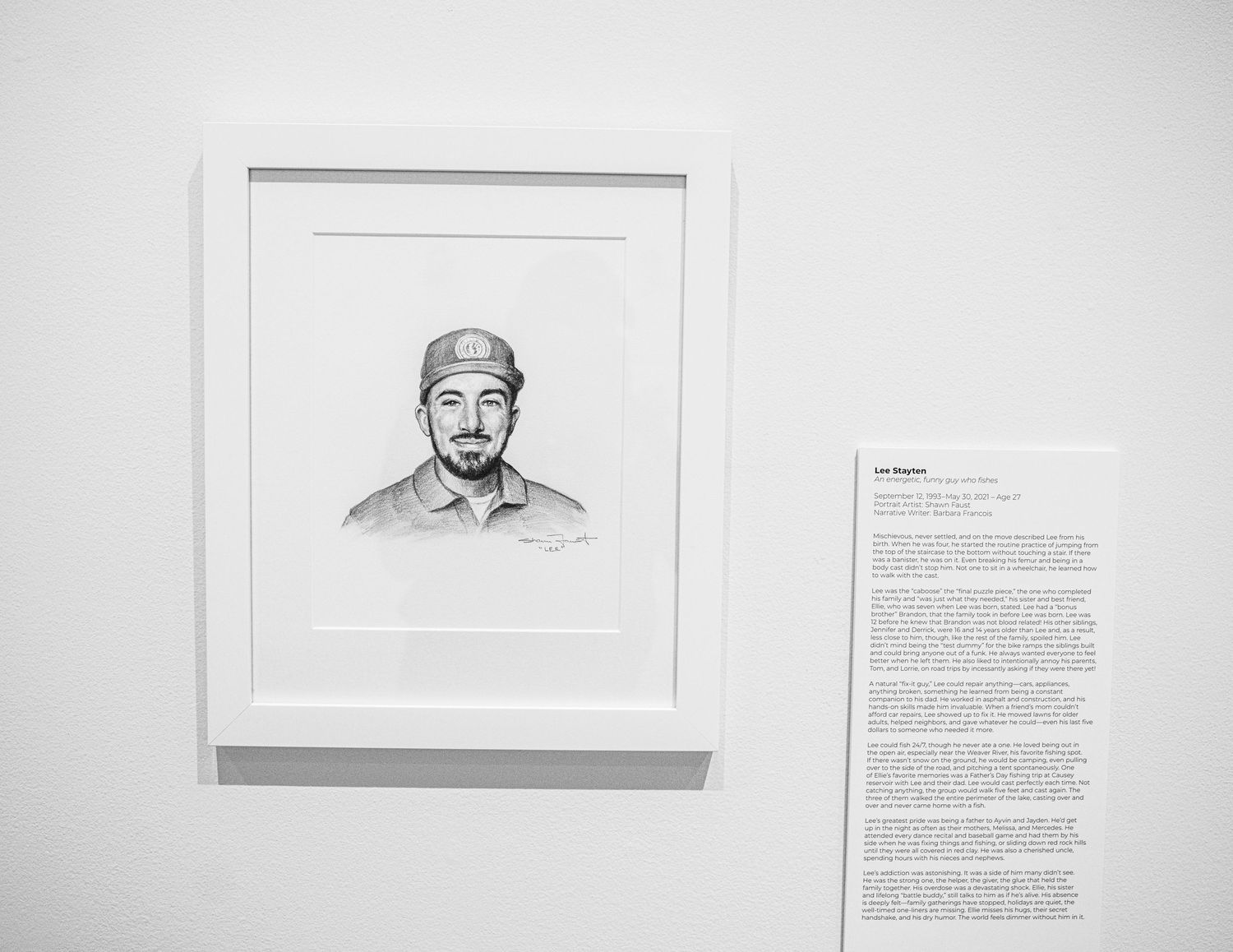 White gallery wall displaying portrait drawings with descriptive text panels mounted beside them.
