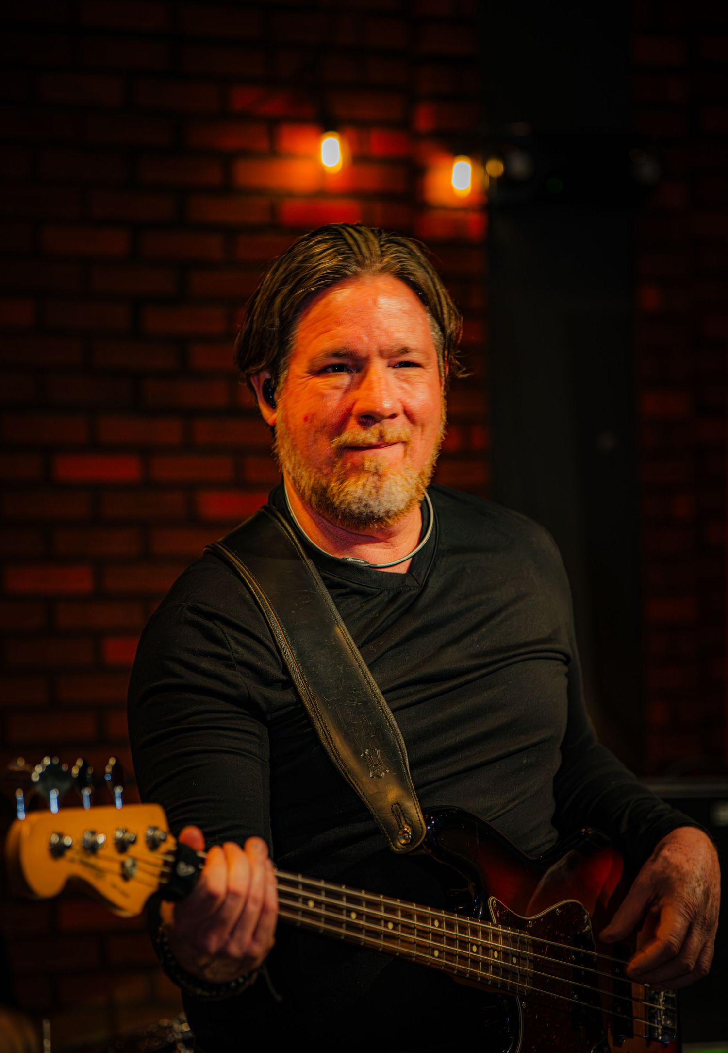 Bass guitarist performing on stage with warm lighting and brick wall background.