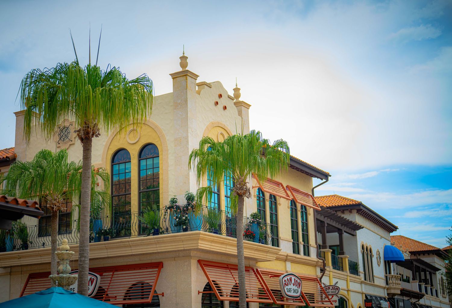 Colorful Mediterranean-style building with arched windows and lush palm trees under a bright blue sky in a vibrant town square.