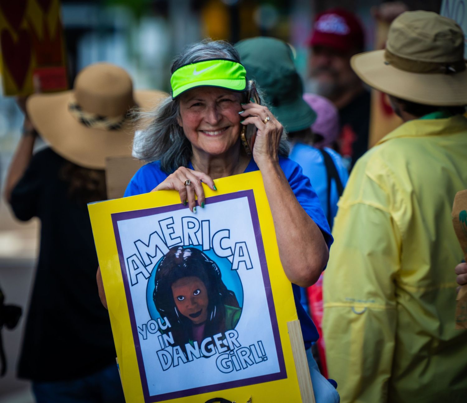 A protester in a neon visor holds up a colorful 'America Dance Club' sign while talking on a cell phone at a rally.