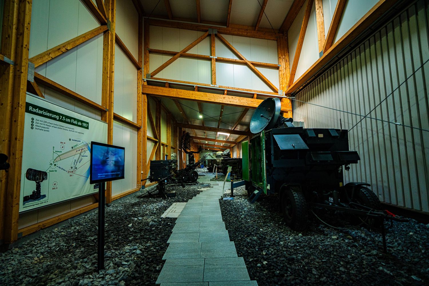 Wooden-beamed museum hall with a stone path leading past vintage mining machinery, an information display screen and a wall map.