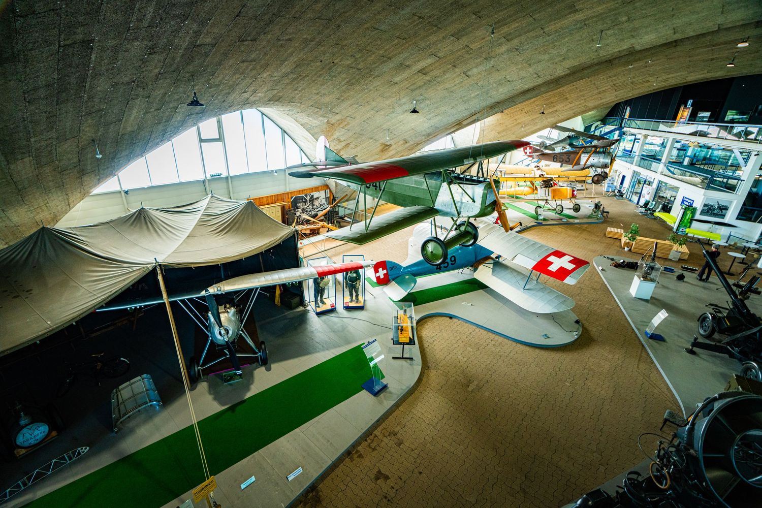 Vintage biplane with Swiss cross markings displayed among historic aircraft in a bright aviation museum hall with a curved ceiling.