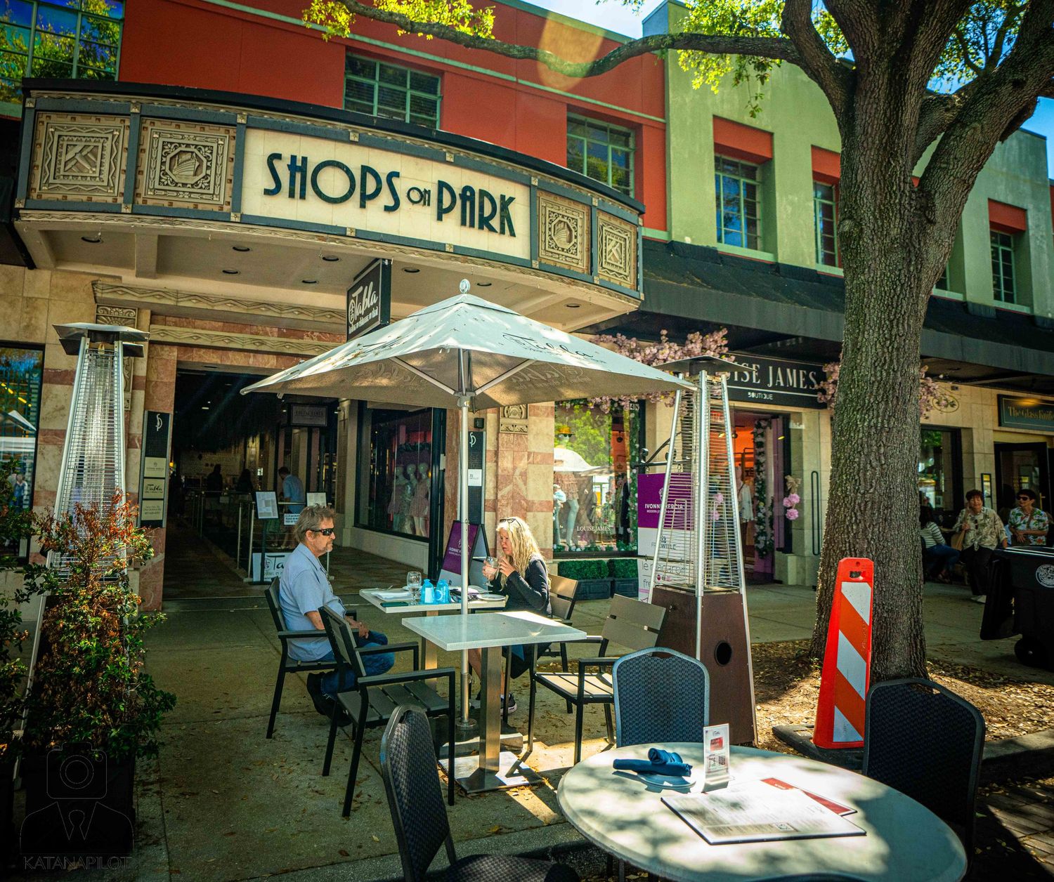 A vibrant street scene showing storefronts and pedestrians walking along a tree-lined urban sidewalk during daytime.