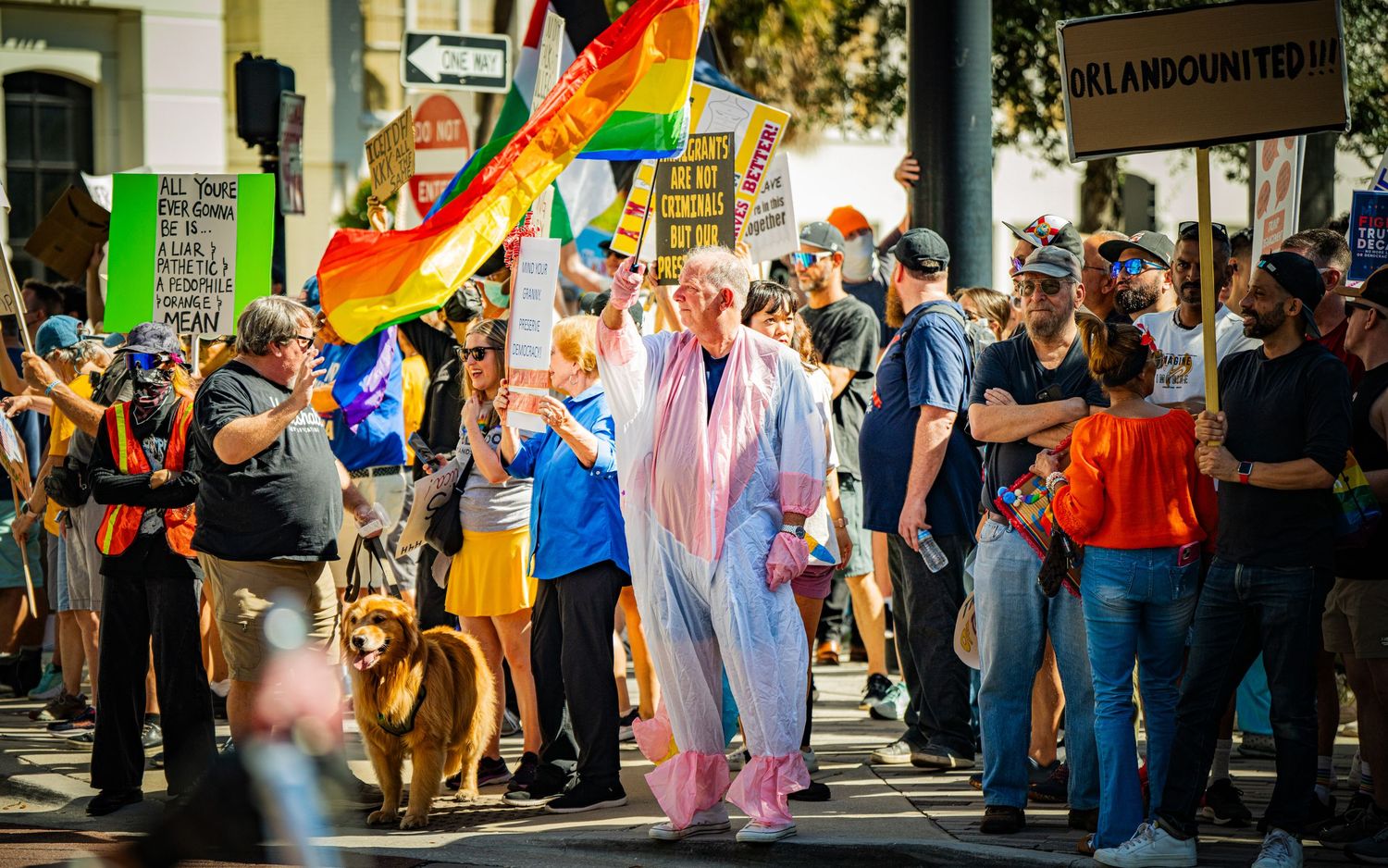Protesters march with rainbow flags and signs while a dog joins the demonstration.