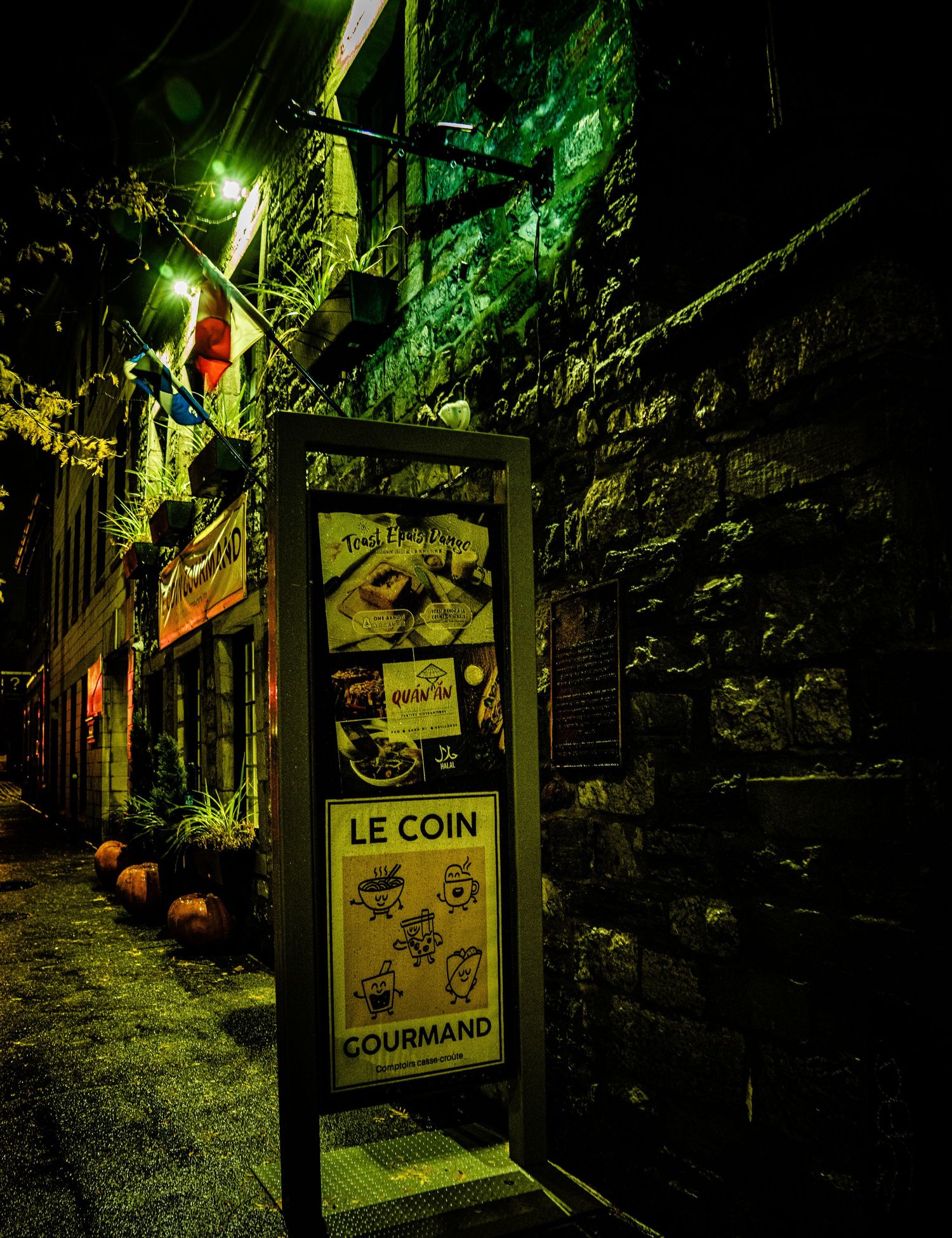 A dimly lit storefront sign reading 'Le Coin Gourmand' illuminated by green and yellow lights against a stone wall at night.