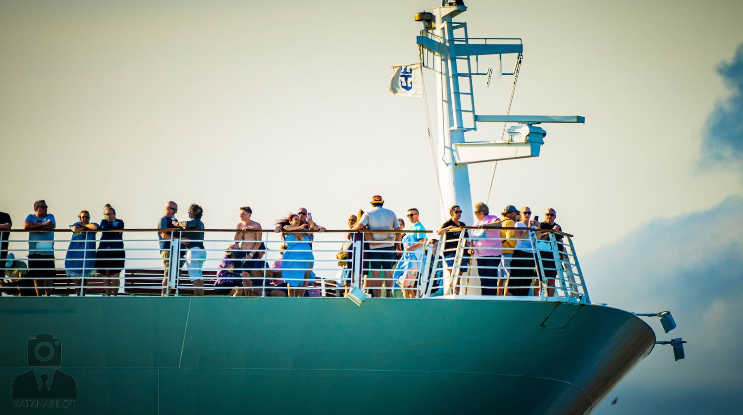 Passengers line the railings of a cruise ship deck against a bright blue sky and ocean backdrop.