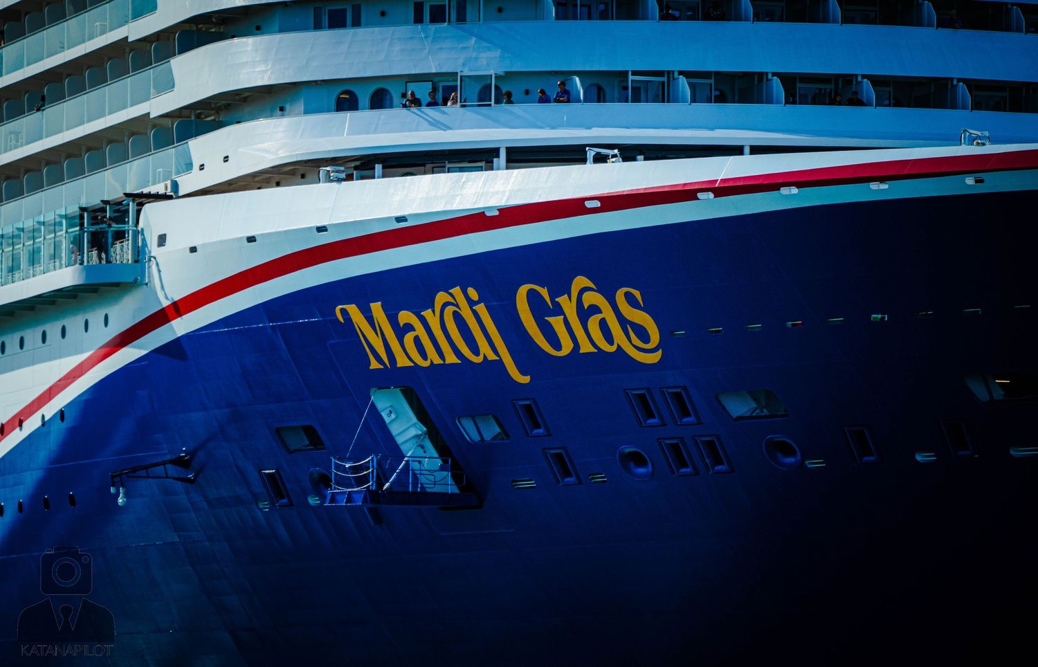 Cruise ship named Mardi Gras docked at port with blue sky and water in background.