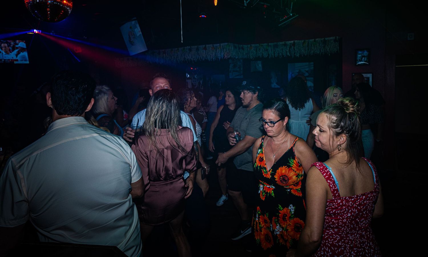 Dimly lit nightclub atmosphere with red and blue accent lighting and dancing silhouettes.