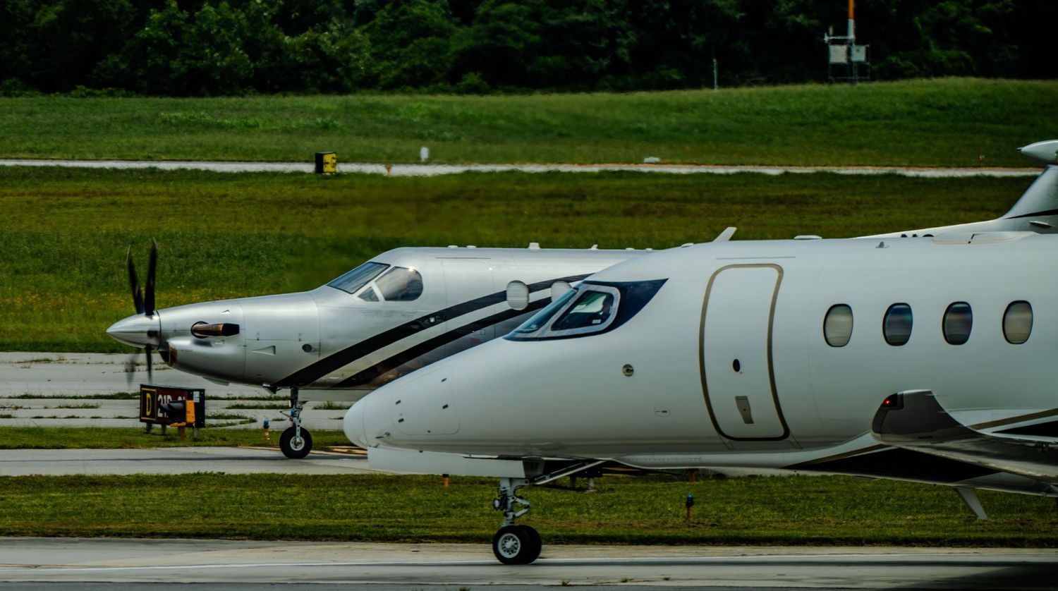 Two white private turboprop aircraft taxiing on a runway surrounded by lush green grass on a cloudy day.