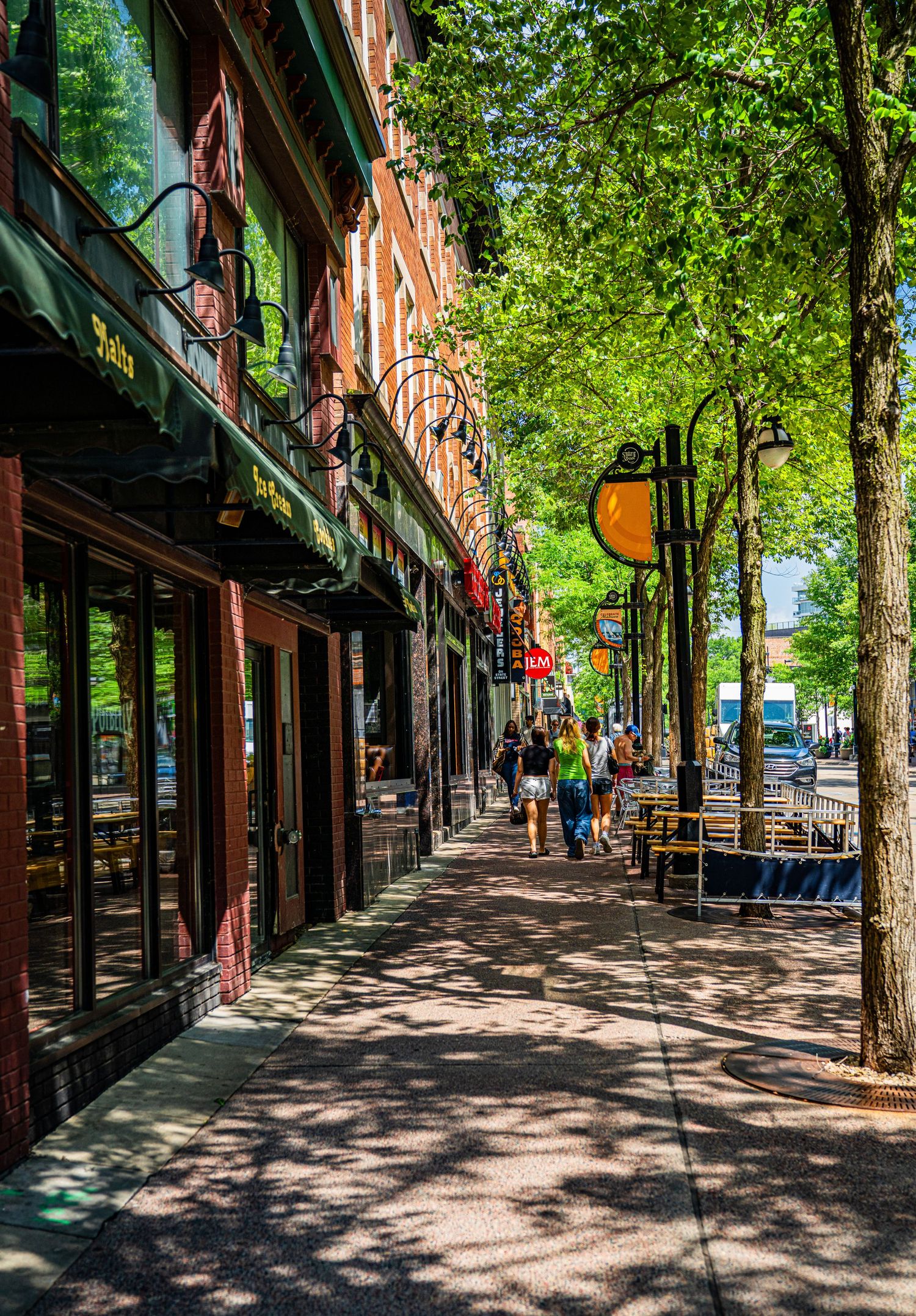 Historic brick storefronts line a shaded sidewalk with trees creating dappled sunlight patterns.