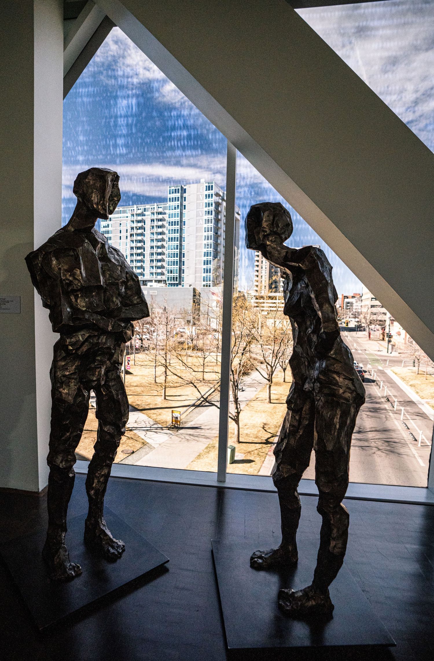 Bronze sculptures of people leaning and looking out windows create a dramatic silhouette against a cityscape view.