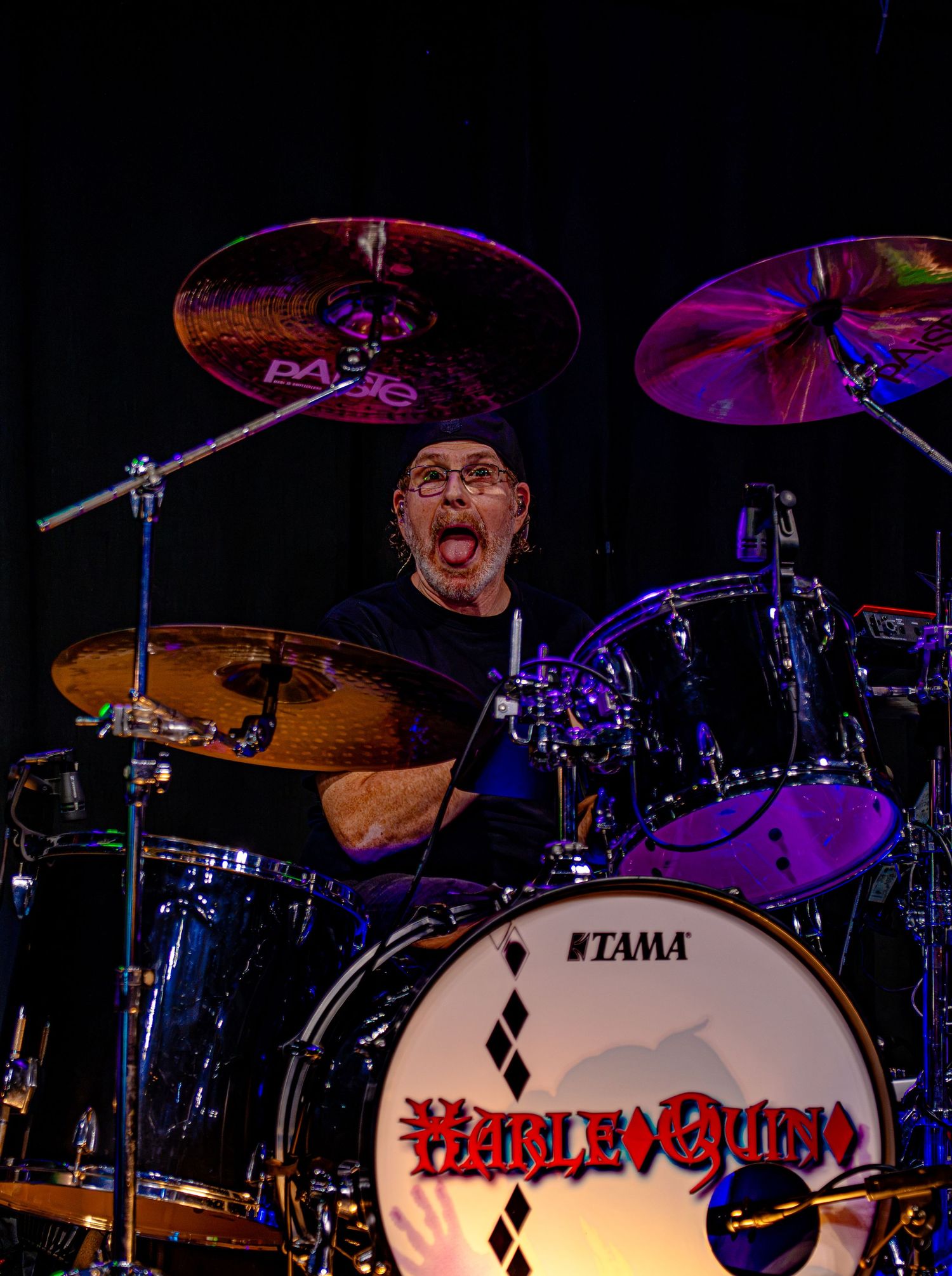 Energetic drummer performing live on a Tama drum kit for the band Harlequin, surrounded by Paiste cymbals on a dark stage.