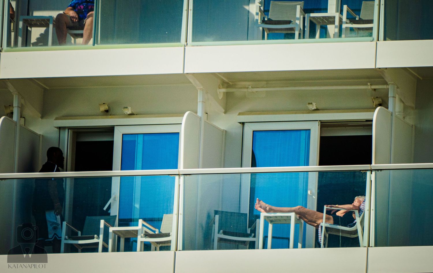 Passengers lounging on private balconies of a modern cruise ship with blue-tinted windows.