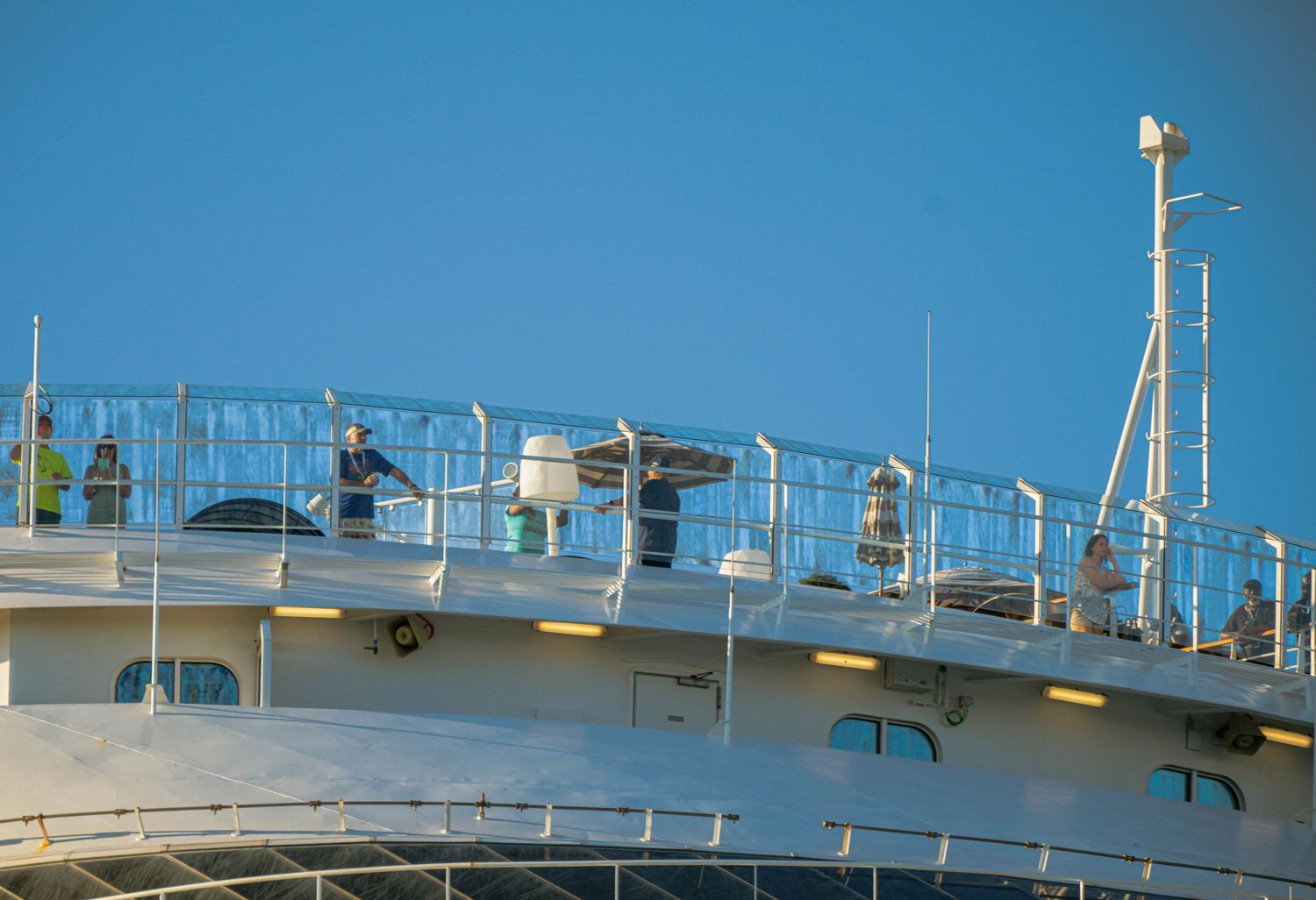 A white cruise ship deck with railings and safety equipment against a bright blue sky background.