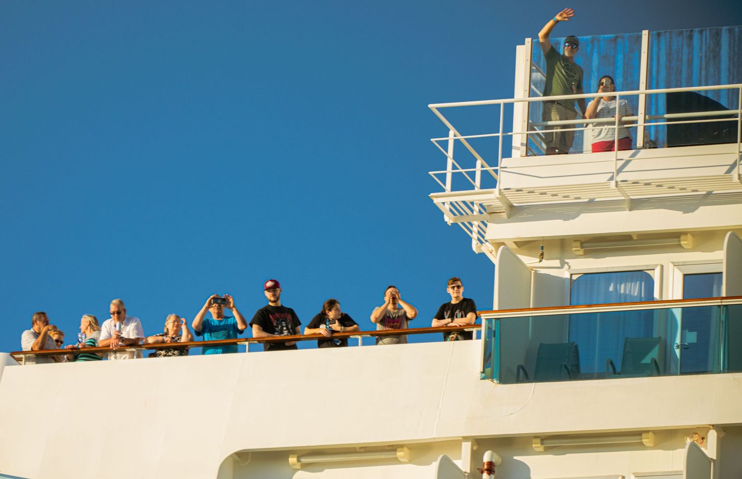 Passengers gather on multiple decks of a white cruise ship against a bright blue sky and ocean background.