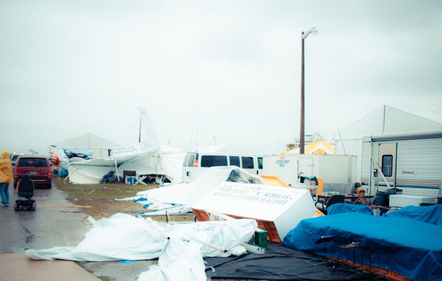 Small aircraft and vehicles parked at an airfield under cloudy skies with tarps covering some equipment.