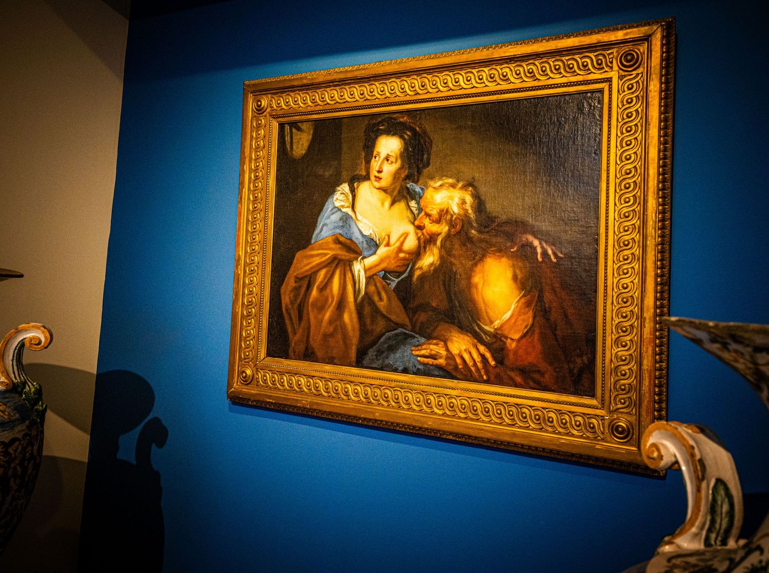 Museum gallery wall with illuminated classical paintings in gilded frames against deep blue background.