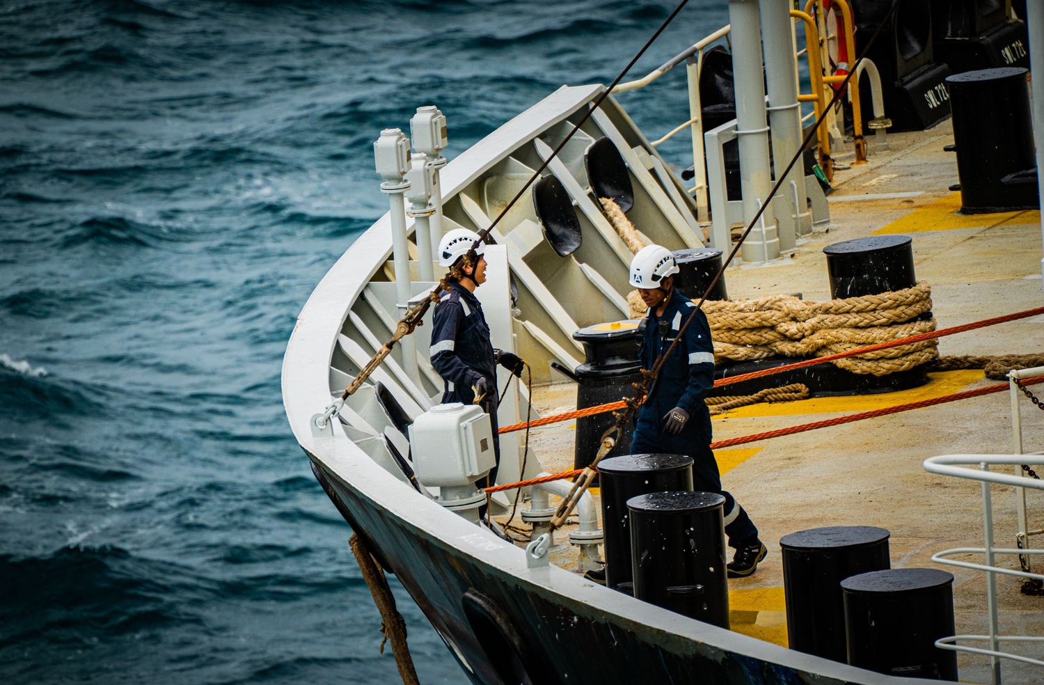 Two sailors in white helmets working on the bow of a ship at sea.