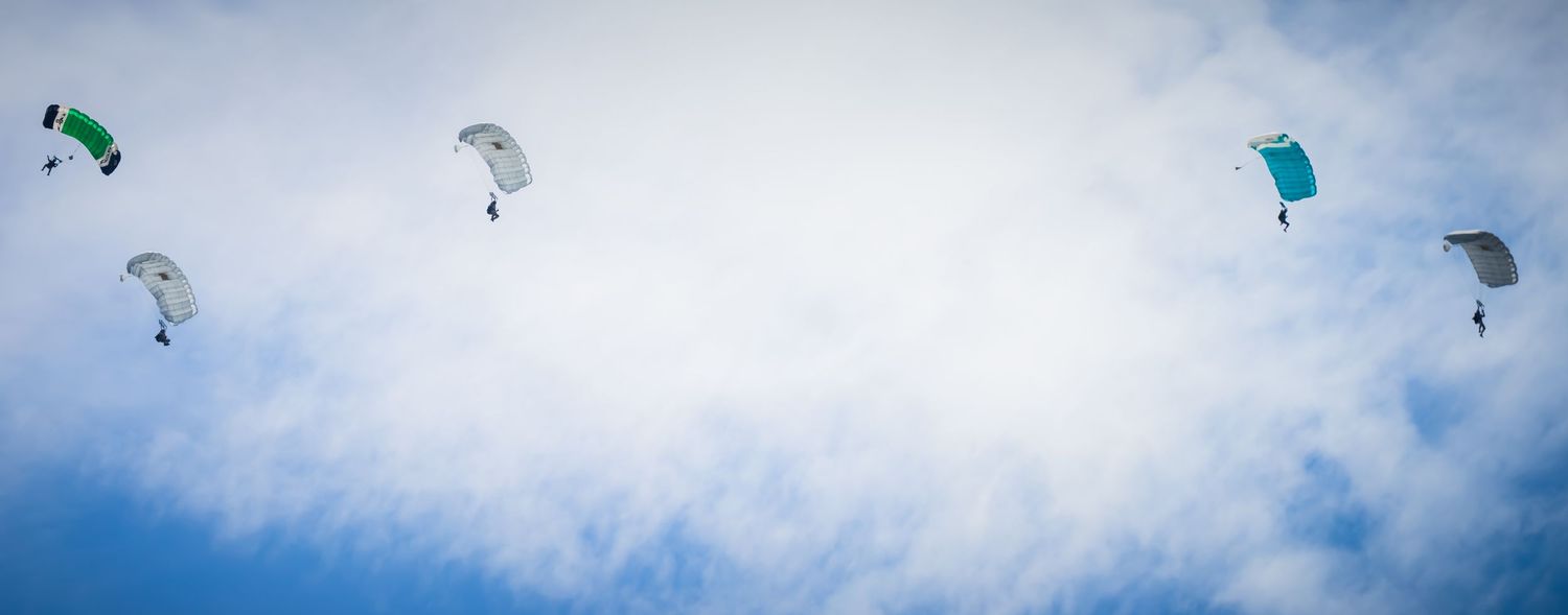 Multiple skydivers with colorful parachutes descend through a bright blue sky with scattered clouds.