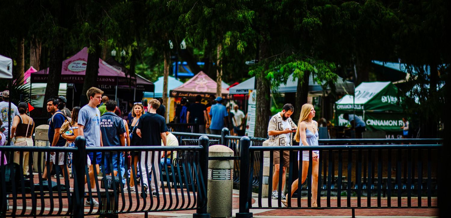 Crowd of people enjoying an outdoor festival market with colorful vendor tents and lush trees in the background.