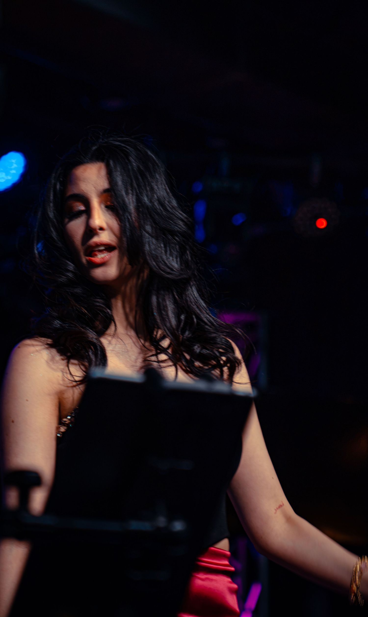 Young woman with long dark hair performing on stage in a black dress under colorful concert lighting.