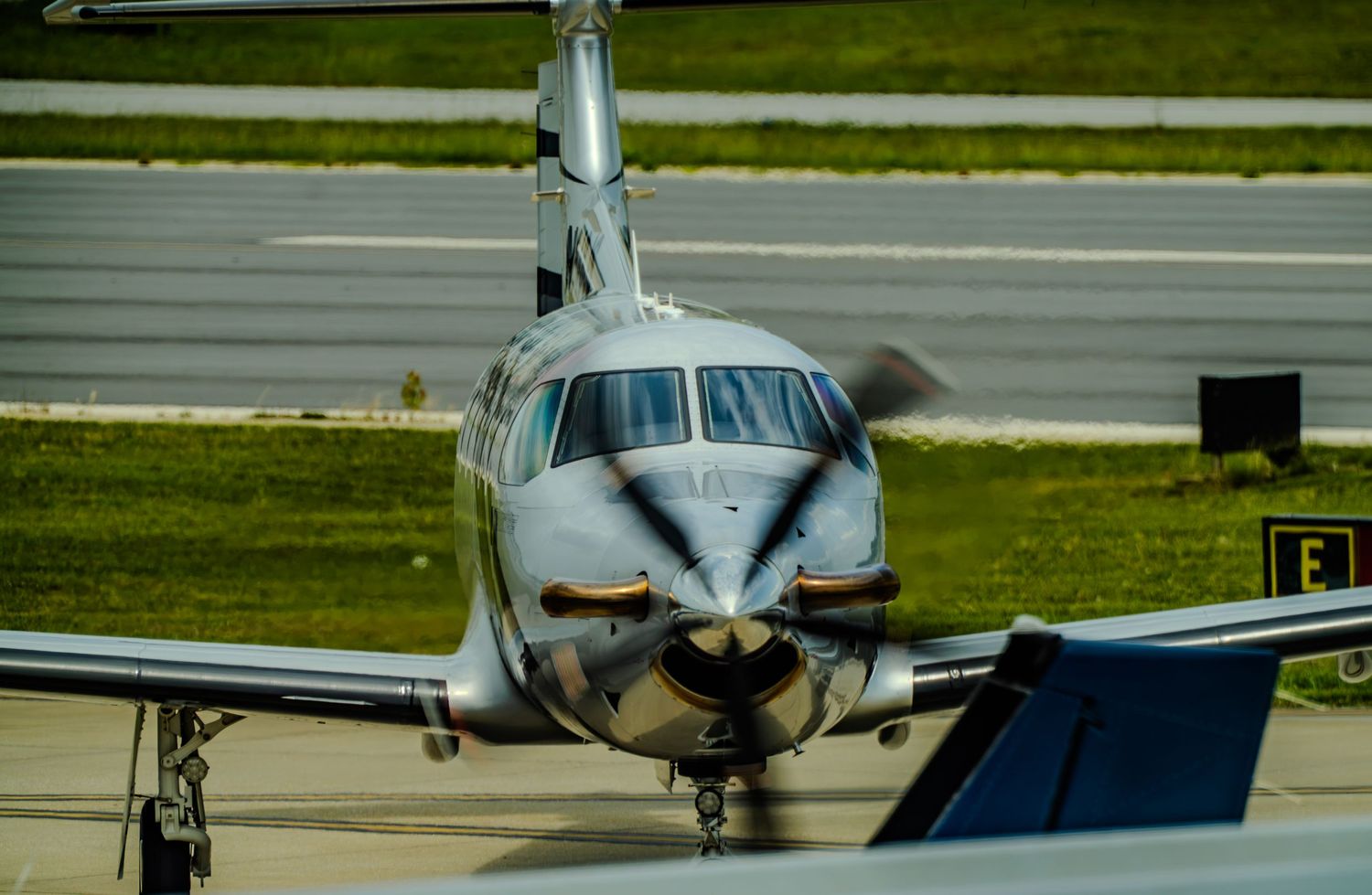 Silver twin-engine propeller aircraft taxiing on runway with spinning propellers at a busy airport.