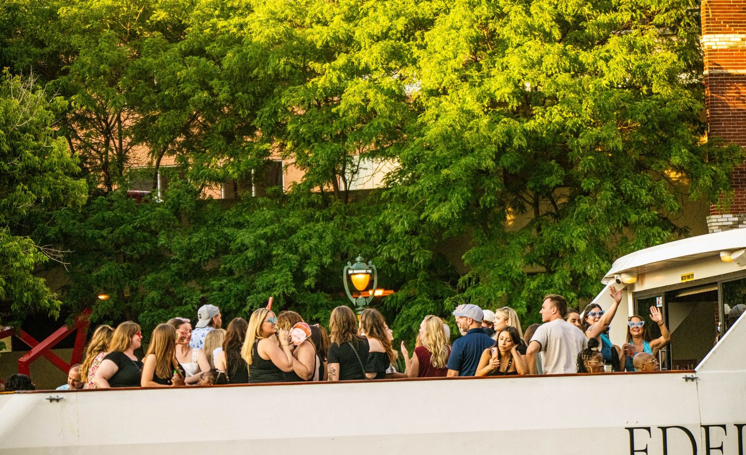 Crowd enjoying summer evening along tree-lined waterfront path.