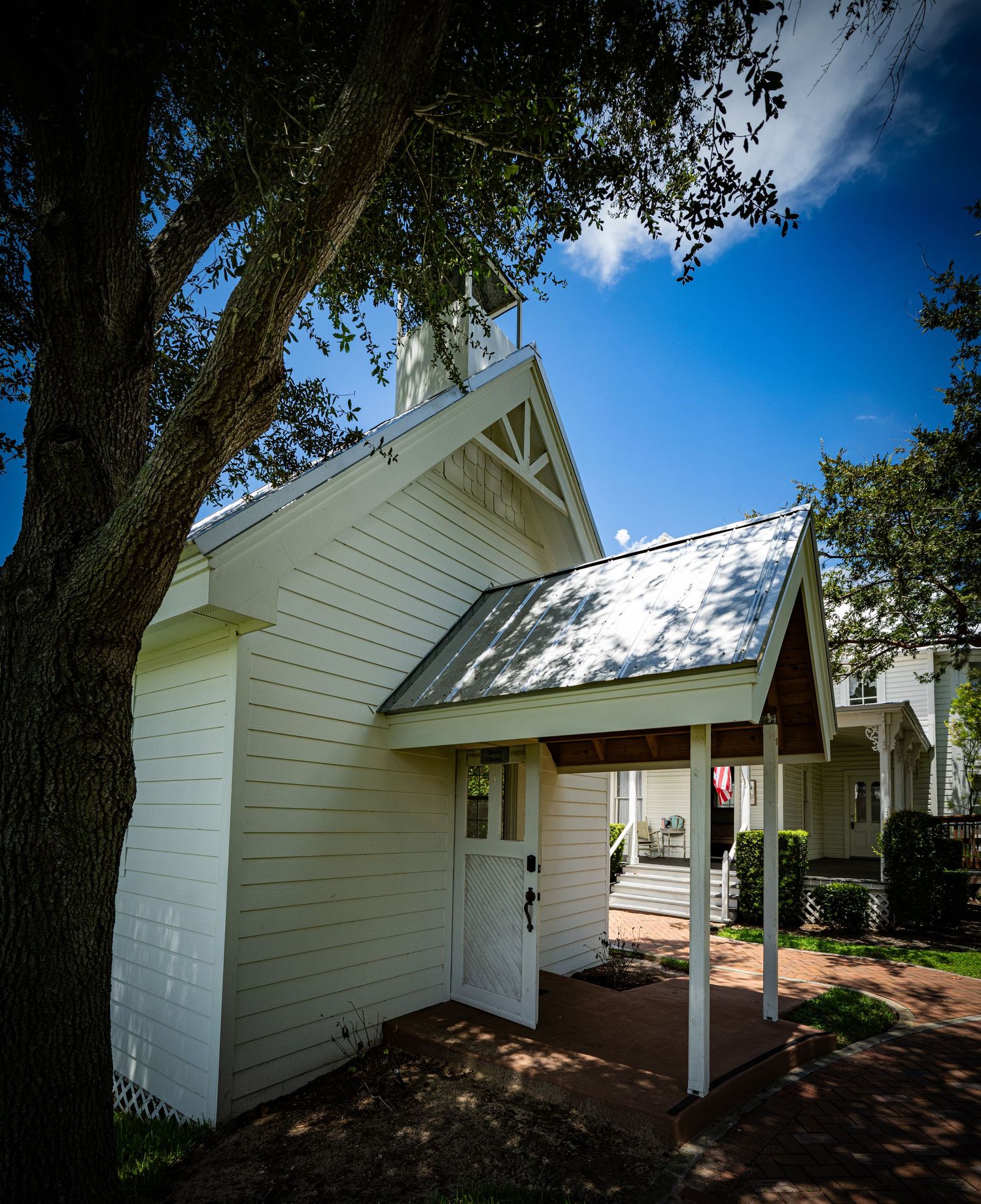 Small white chapel with a steeple and covered entrance framed by a large tree against a bright blue sky.