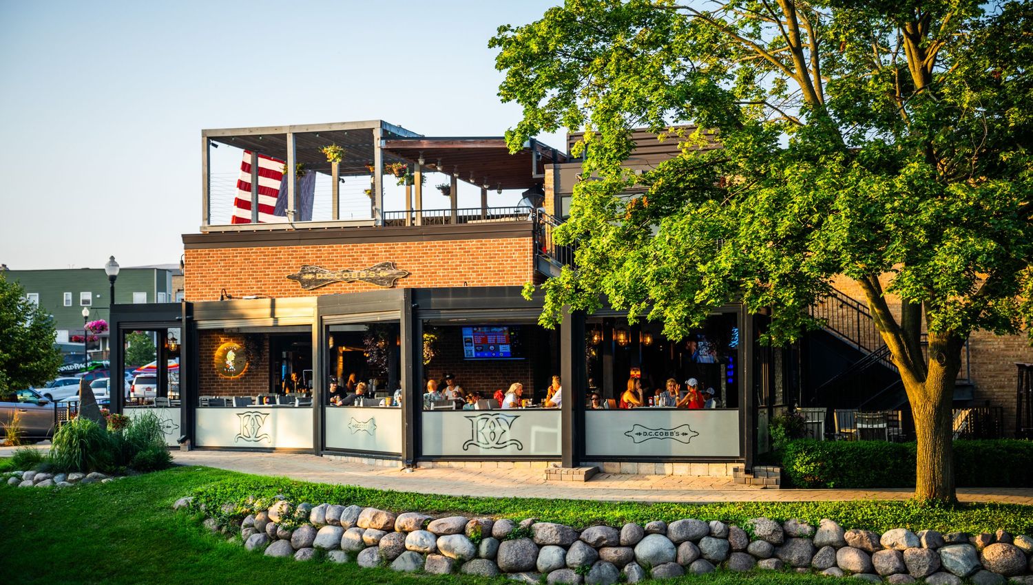 Riverside restaurant with outdoor upper deck seating area surrounded by green trees.