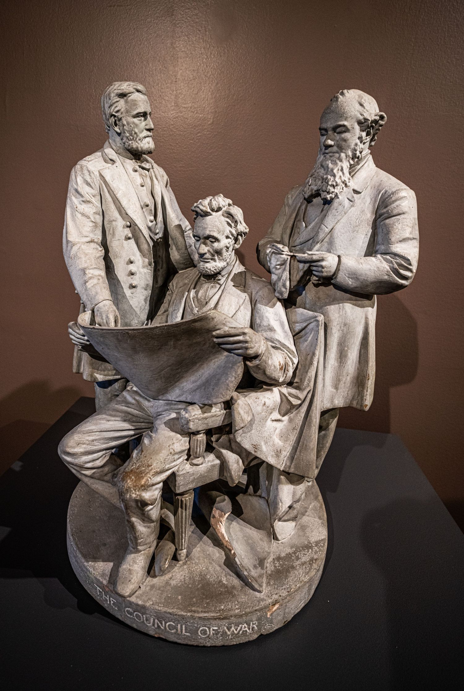 Historical sculpture depicting seated figures in formal attire, displayed on dark pedestal.