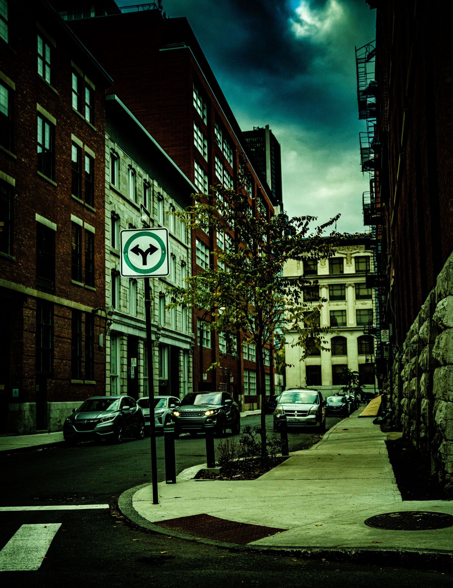 Dark urban street with brick buildings, parked cars, and trees under an ominous teal-colored stormy sky.