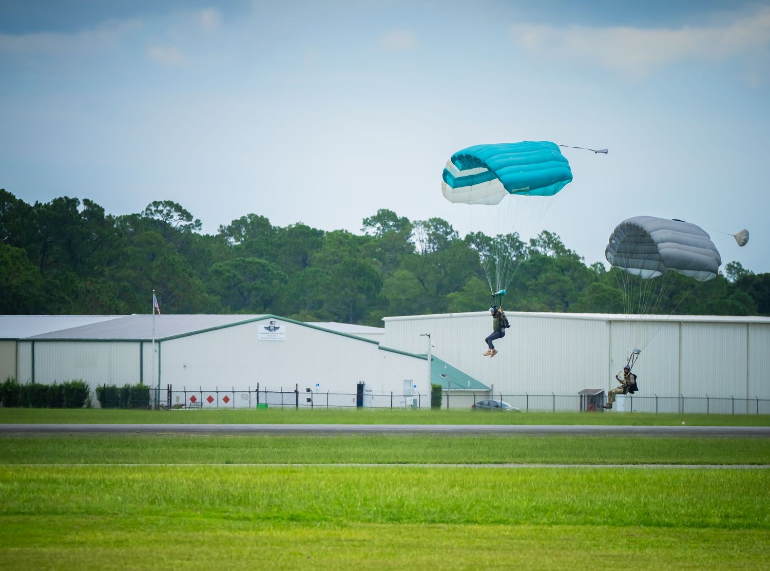 Skydiver approaching landing zone with blue parachute against cloudy sky and buildings.