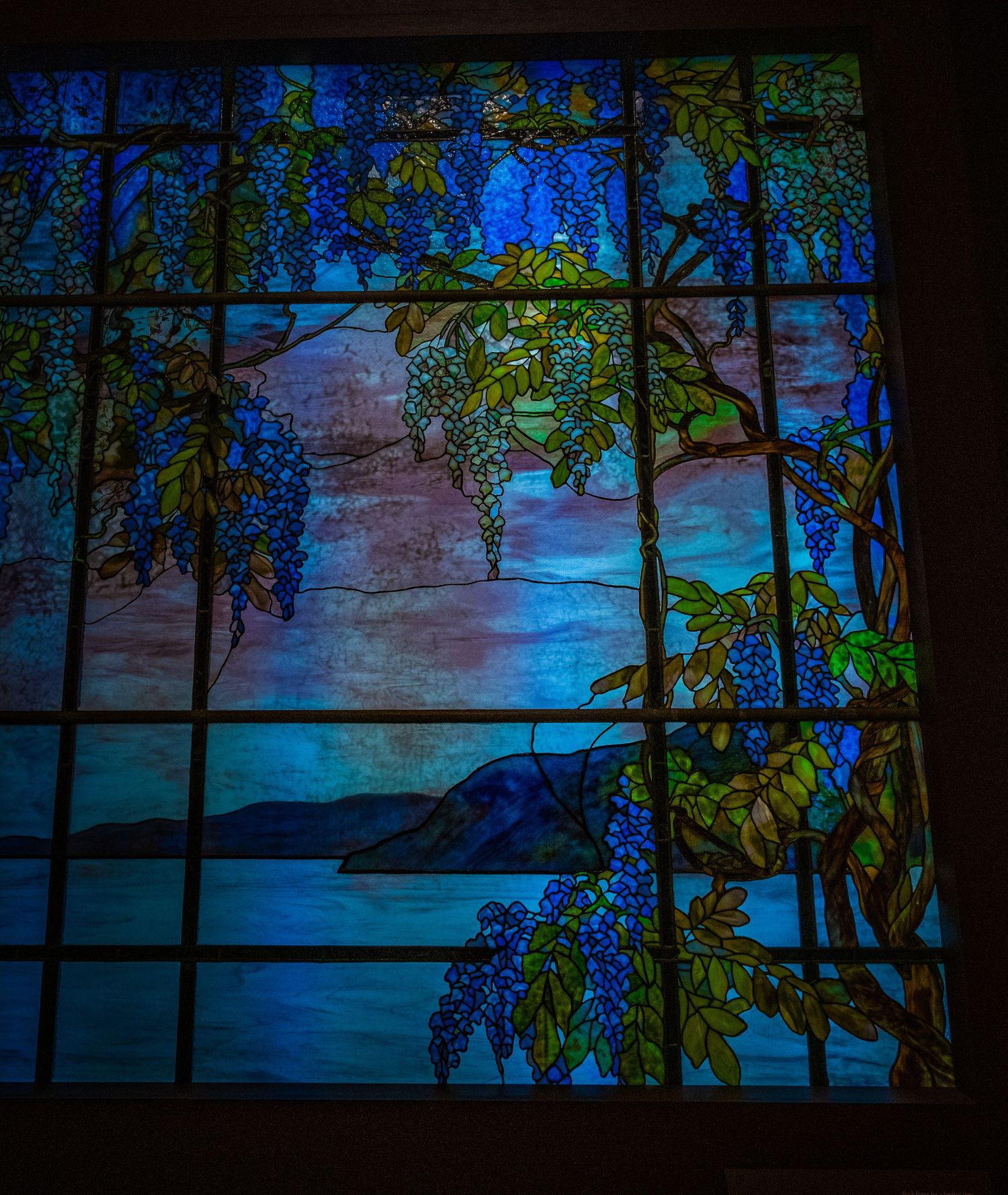 Stained glass landscape scene with hanging wisteria vines framing a serene blue waterfront view.