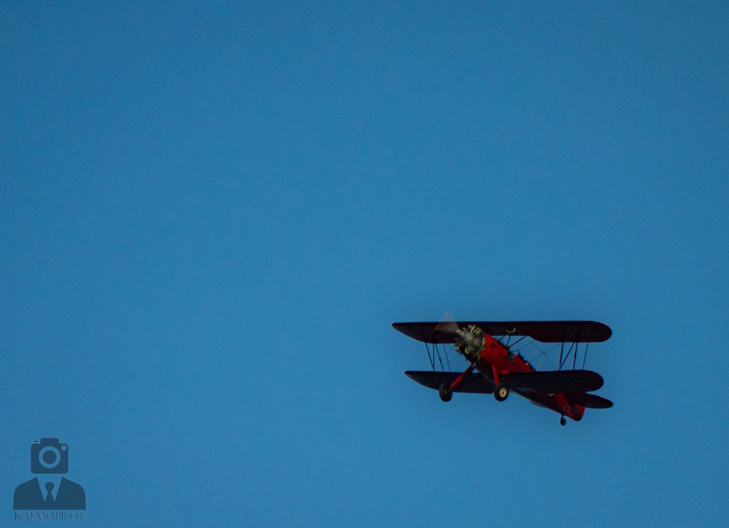 Small silhouette of a biplane flying against a bright blue sky background.