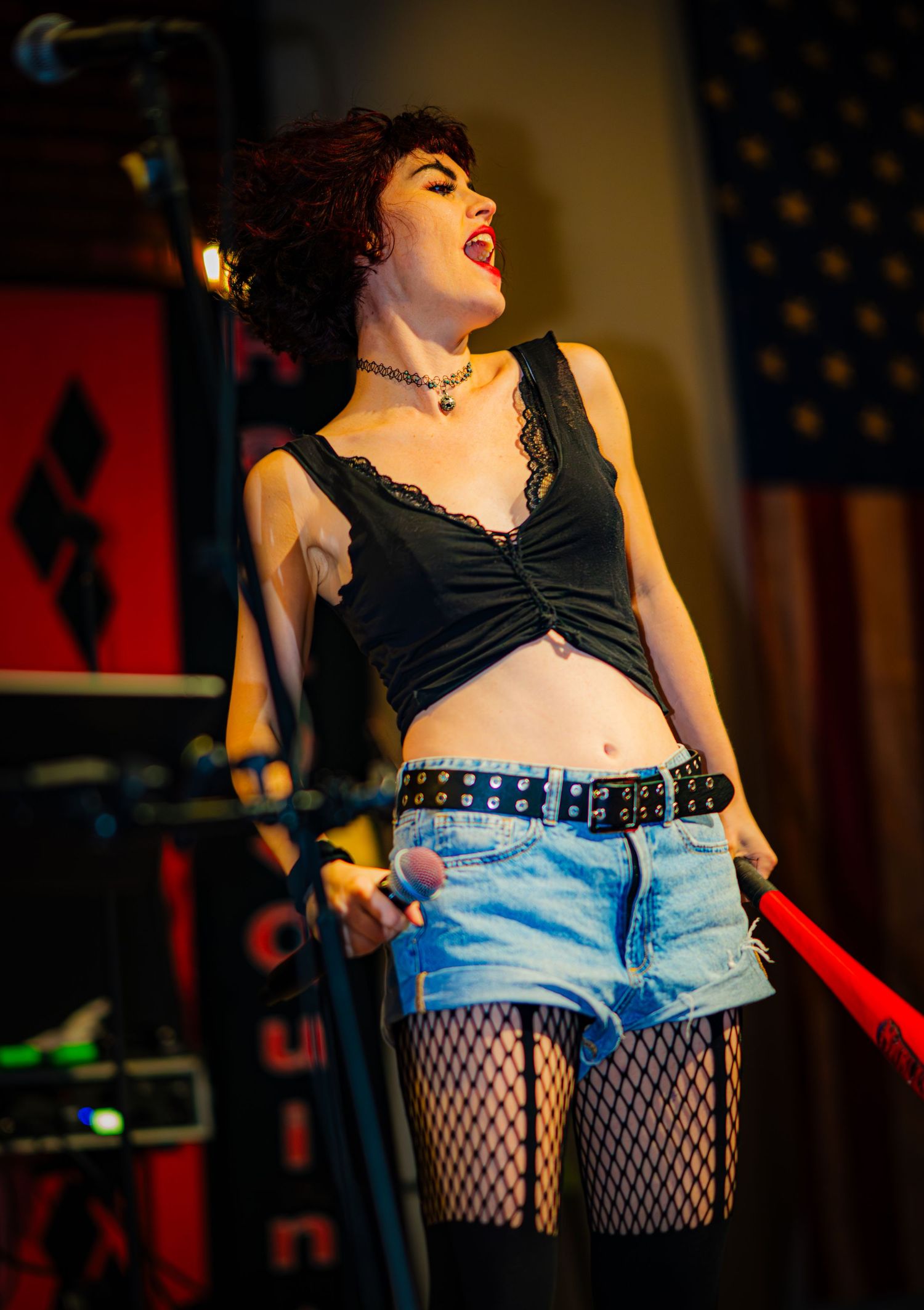 Punk rock performer in fishnets and denim shorts singing passionately on stage with American flag backdrop.