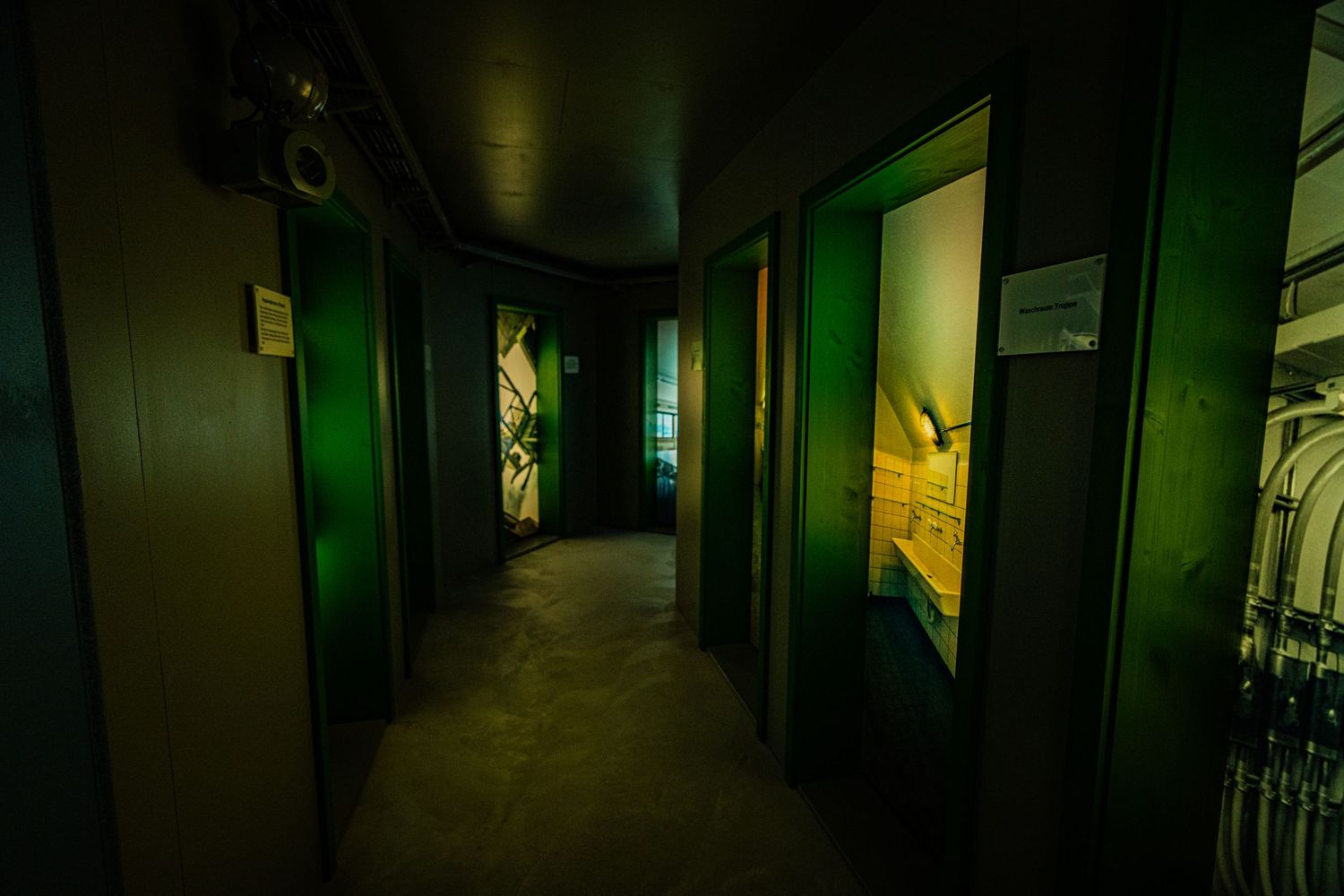 Dimly lit hallway with green lighting leads past open doorways and glass windows, creating a moody, eerie industrial atmosphere.