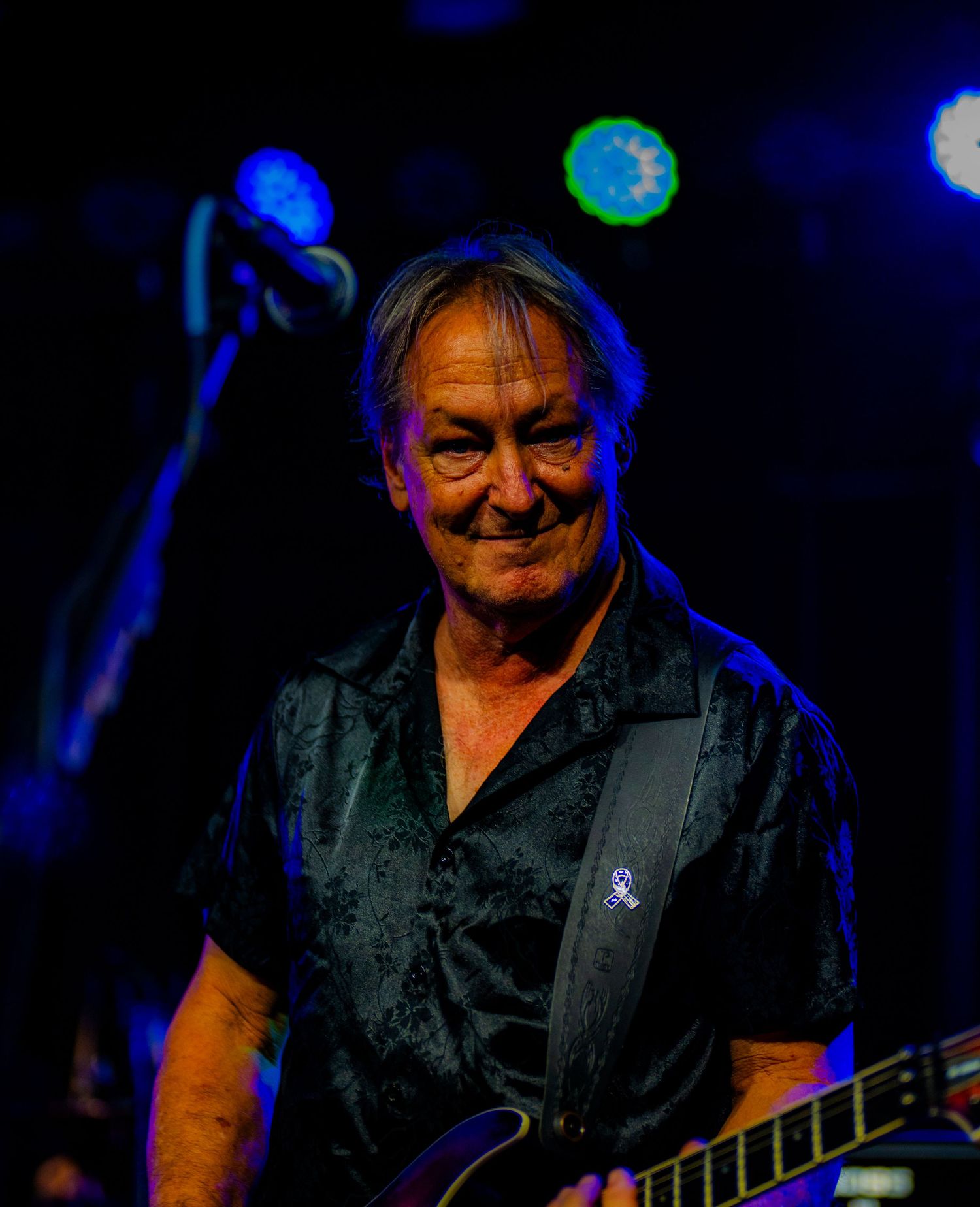Smiling older male guitarist performs on stage under blue concert lights holding an electric guitar.