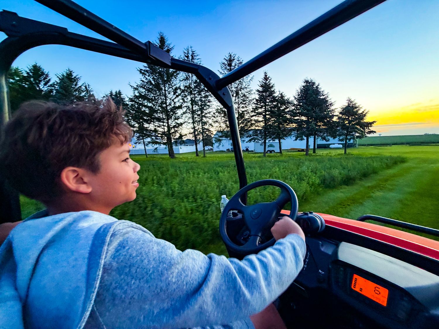 Young boy driving a red UTV golf cart across lush green fields at sunset with tall trees in background.
