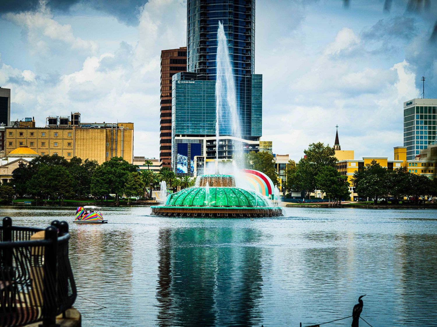 A colorful fountain sprays water on a lake with city buildings in the background.