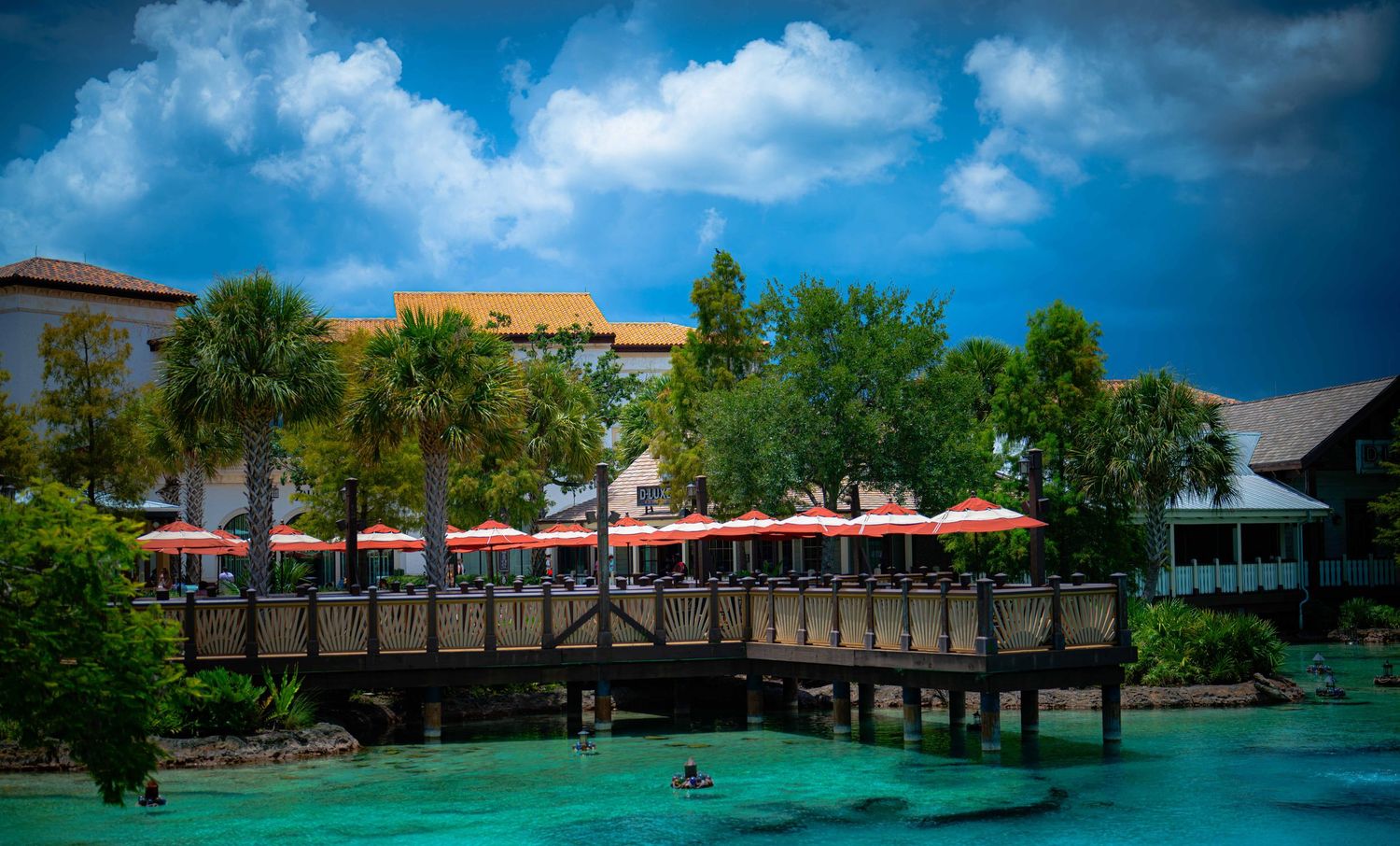 A wooden bridge over turquoise water leads to a tropical resort with red umbrellas and palm trees under dramatic clouds.
