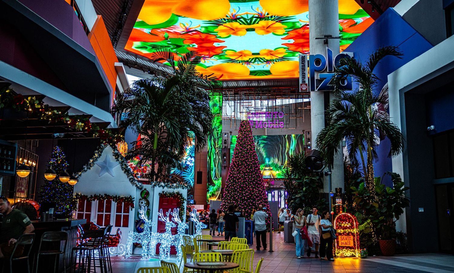 Colorful illuminated shopping mall interior with palm trees and vibrant ceiling decorations creating a festive atmosphere.